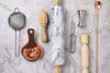 a collection of cocktail tools, strainers, juicer, rolling pin, peeler, jigger, muddler, spoon, against a marble backdrop